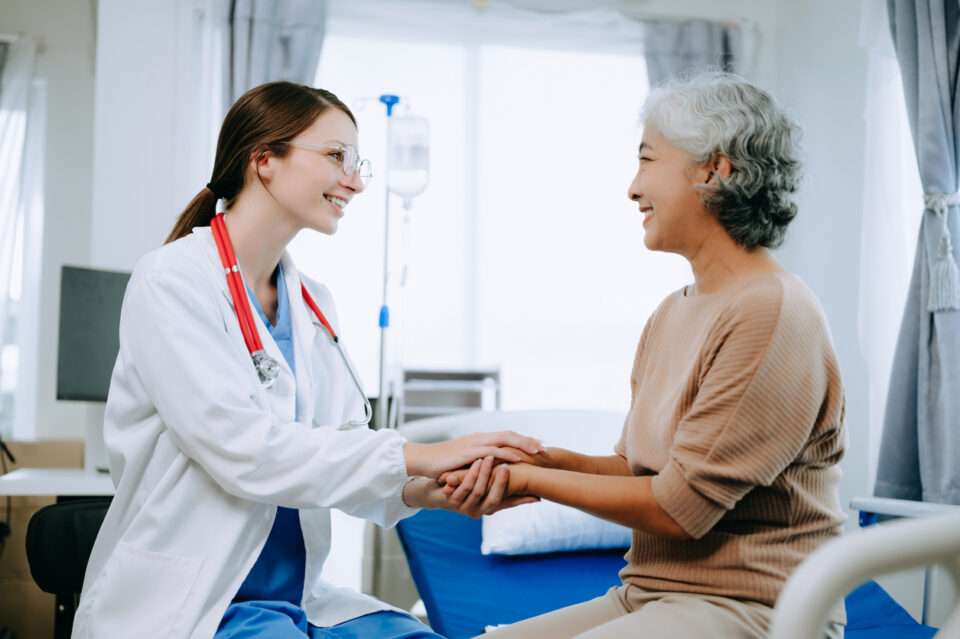 Friendly Female Head Nurse Making Rounds does Checkup on Patient Resting in Bed. She Checks tablet while Man Fully Recovering after Successful Surgery in hospital